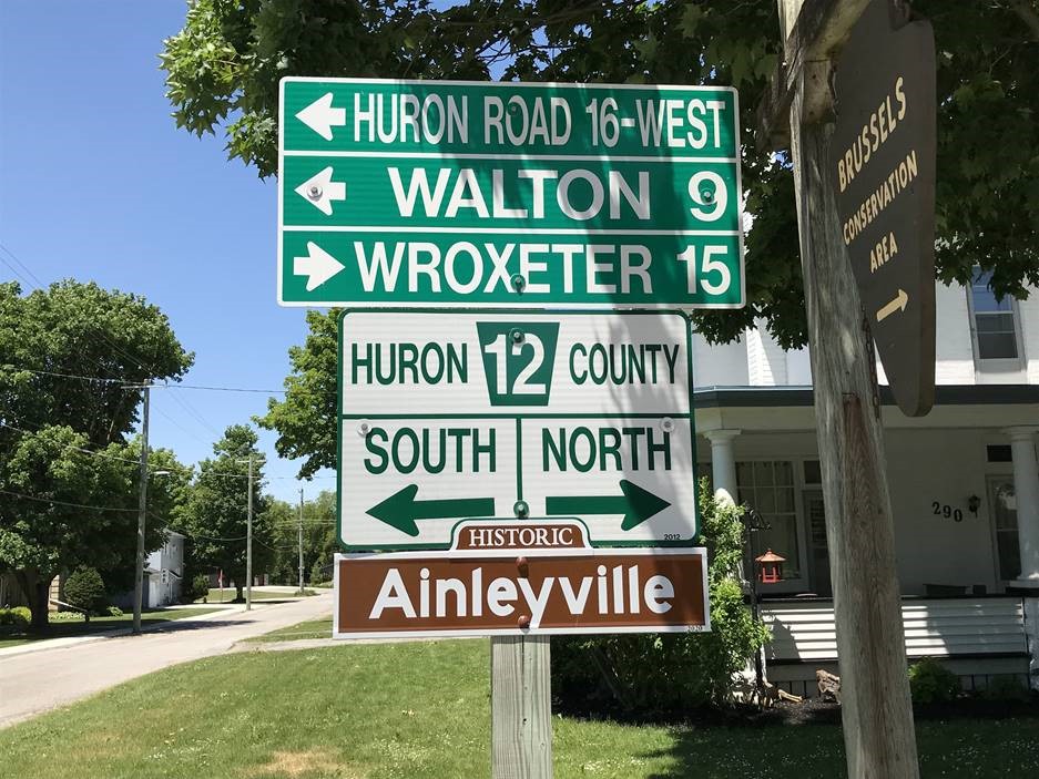 Huron Historic Settlements: Ainleyville highway sign. Brown "Ainleyville" sign below white directional sign for Huron County Road 12 north and south, below green kilometre distance signs for Walton, Wroxeter.