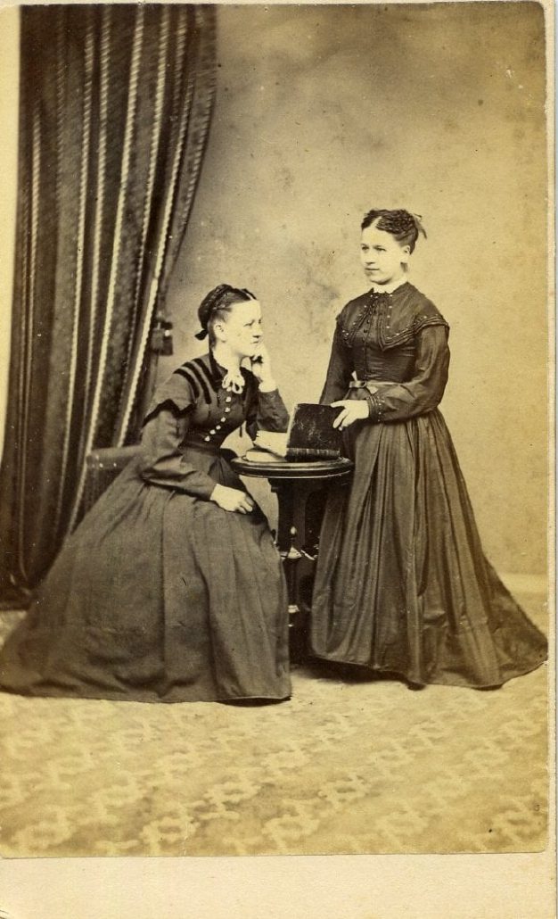 Sepia photograph of two young women in dark dresses, one seated and one standing.