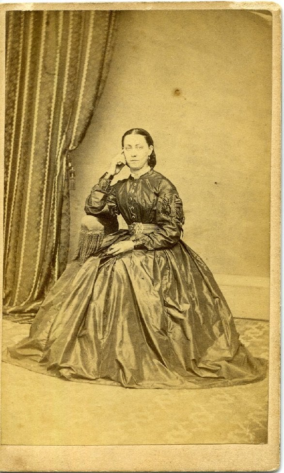 Sepia photograph of seated woman in hoop skirt. Curtain to the left.