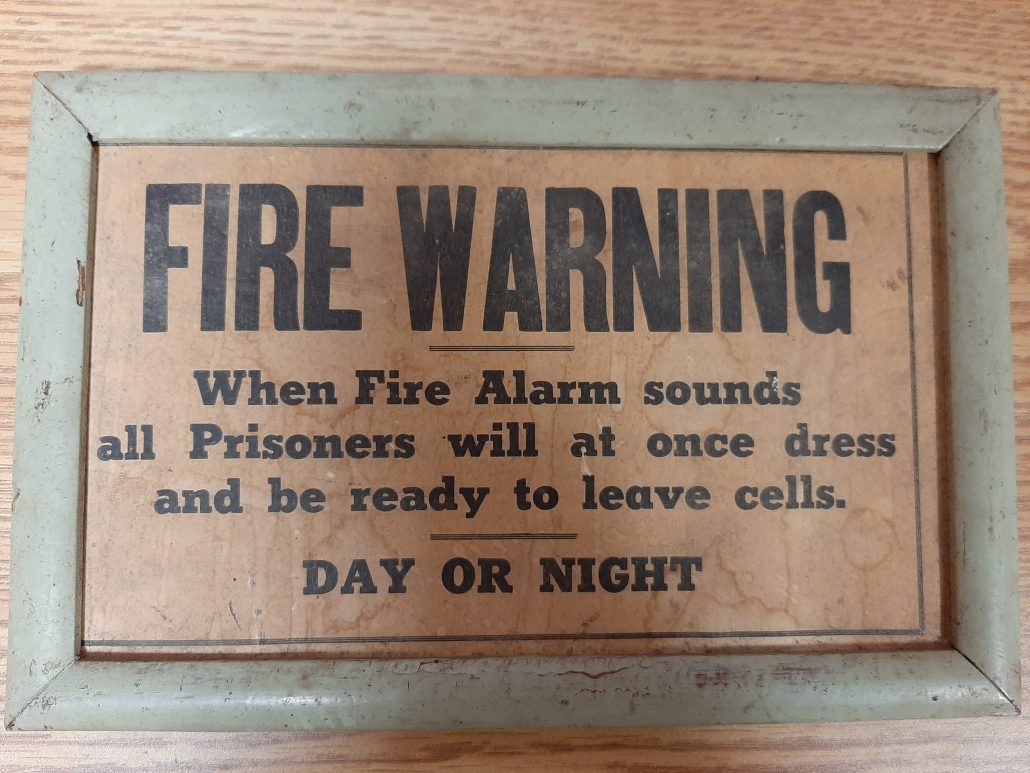 Wooden sign with black text: "Fire Warning: When Fire Alarm sounds Prisoners will at once dress and be ready to leave cells. DAY OR NIGHT."