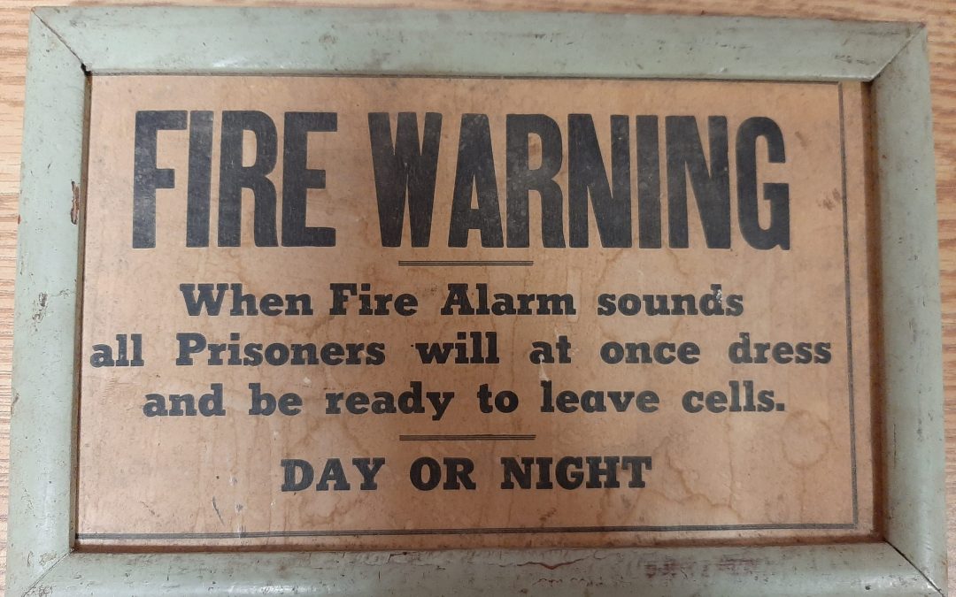 Wooden sign with black text: "Fire Warning: When Fire Alarm sounds Prisoners will at once dress and be ready to leave cells. DAY OR NIGHT."