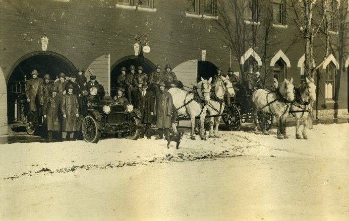 Photograph of firefighters, vehicles and animals in front of fire station.