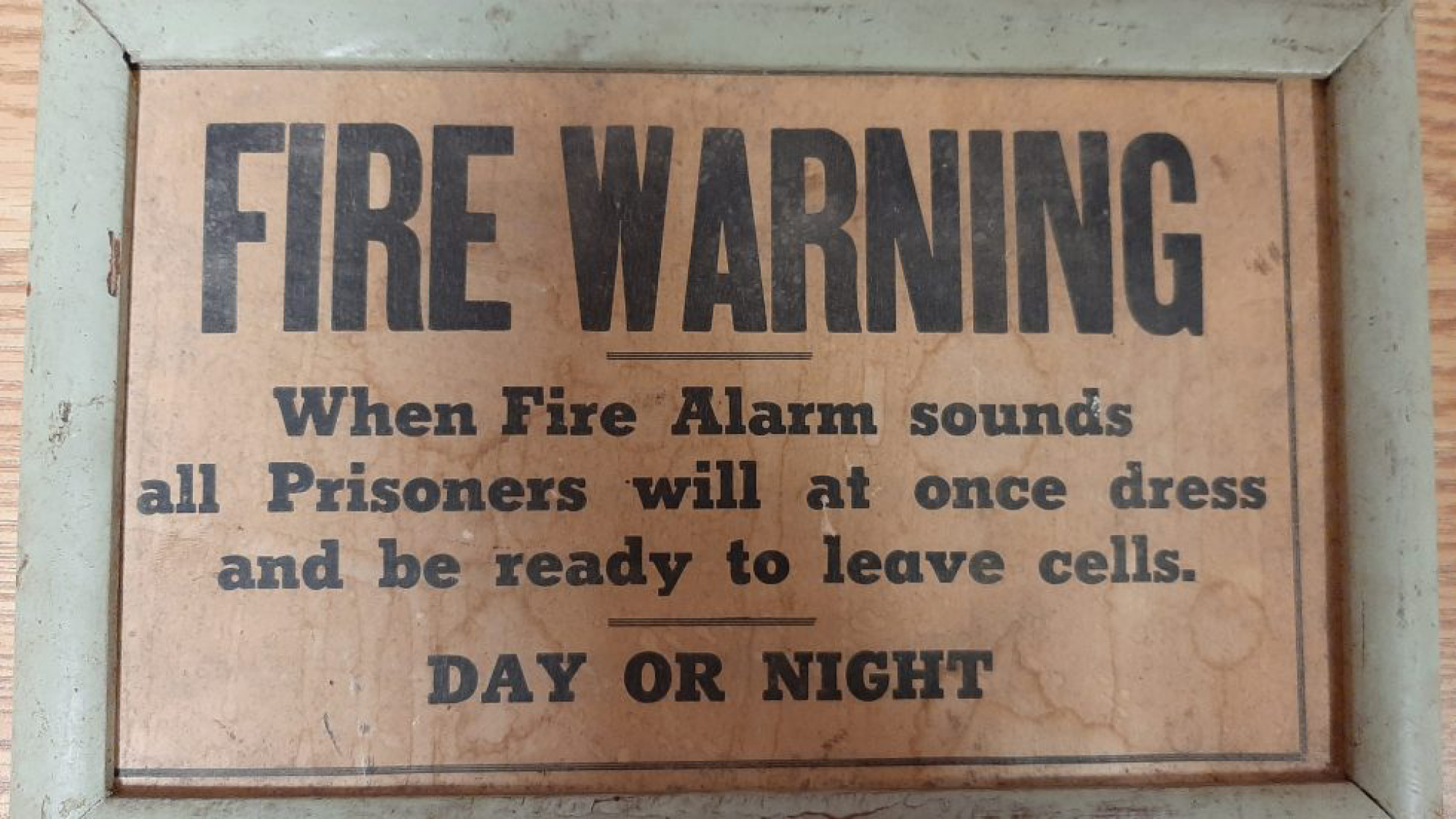 Wooden sign with black text: "Fire Warning: When Fire Alarm sounds Prisoners will at once dress and be ready to leave cells. DAY OR NIGHT."