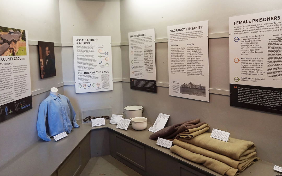 Image of the new exhibit at the Huron Historic Gaol. Features information panels hung on a wall and a variety of Gaol artifacts on a bench