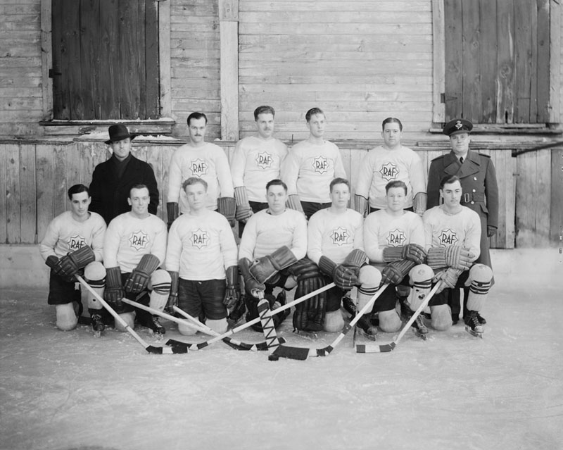 Posed black and white photograph of a hockey team. Eleven male players are wearing white sweaters with the letters "RAF." One man to left in dark coat and hat. One man to right in military uniform.