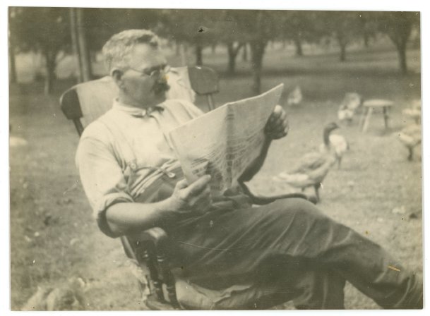 Black and white photograph of a man seated on a chair outdoors reading a newspaper. He has light hair, a dark moustache and is wearing glasses. In the background are geese.