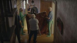 Photograph of multiple people, all senior citizens, holding a rug in an indoor setting, and one person in front with a camera. 
