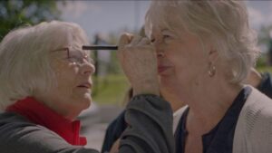 Close-up photograph of a woman with white hair and waring glasses applying make-up with an eyeliner pencil to another woman with white hair. 