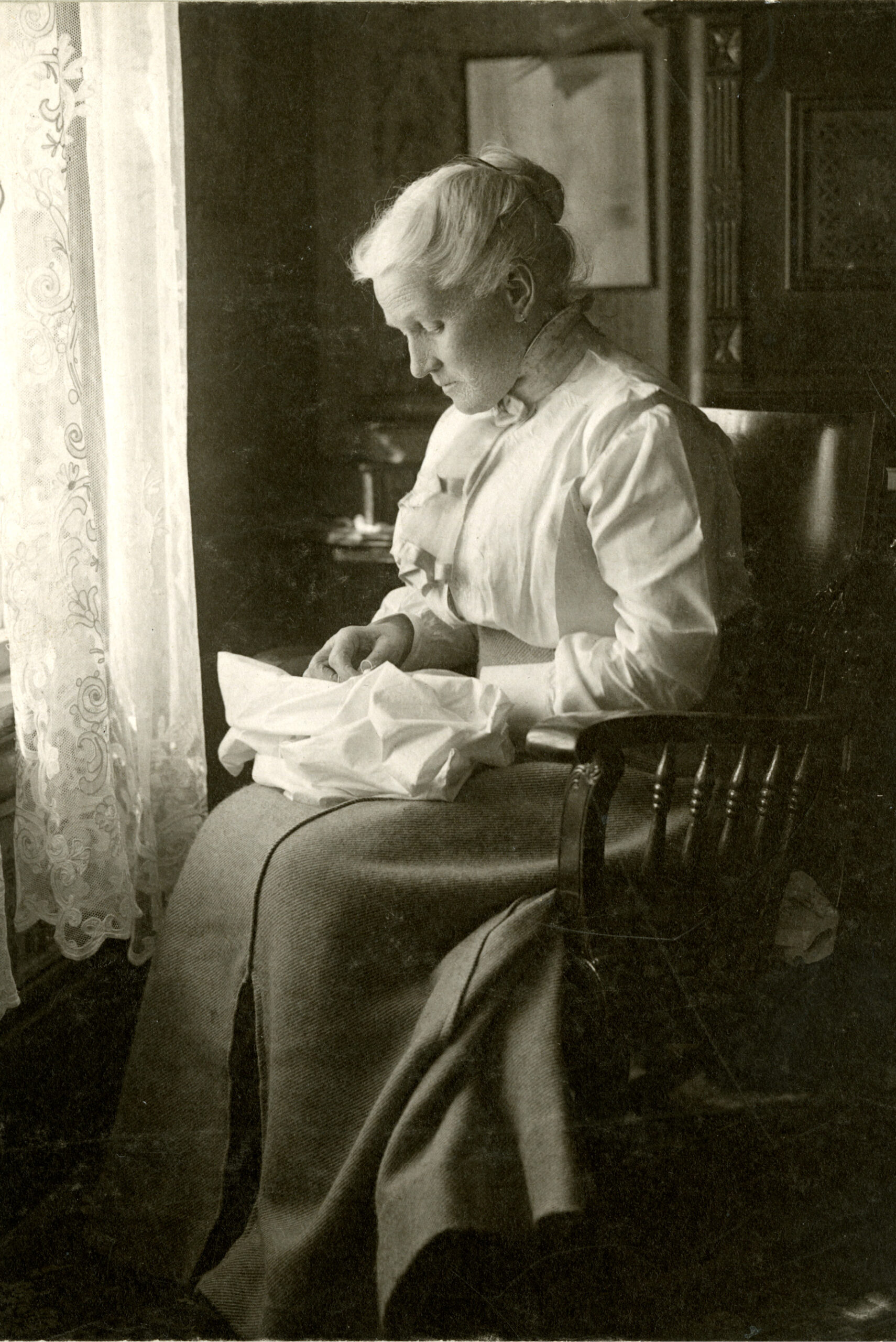 Historic black and white image of an elderly woman sitting in a chair sewing